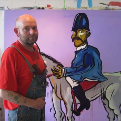 Artist in a red shirt standing in front of a painting of a rider on his horse